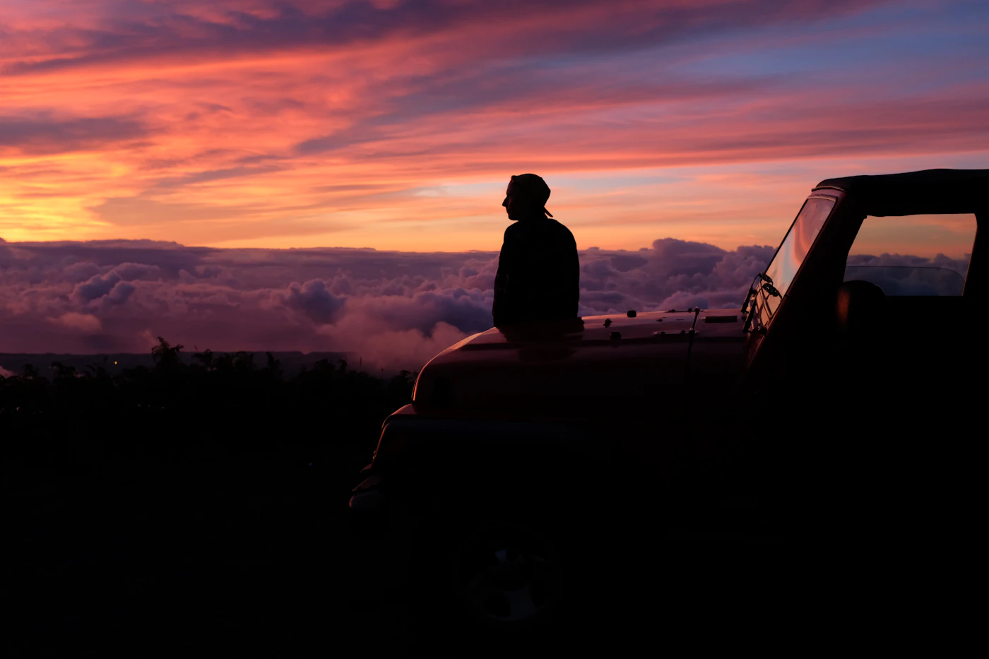 A silhouette sitting on a car hood watching the sky at dusk